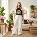 Cheerful woman in black tee with cream cardigan, white pants and colorful accessories in bright airy living room with green plants, pink flowers and black accents